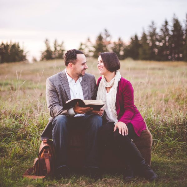Photo of a smiling couple sitting in a field with the Bible between them held by the husband, as if they are reading it together.
